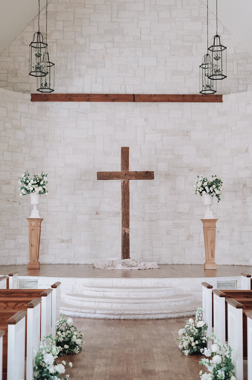 A bare barn wood cross centered beneath a wooden beam between two pinewood pedestals with floral arrangements.