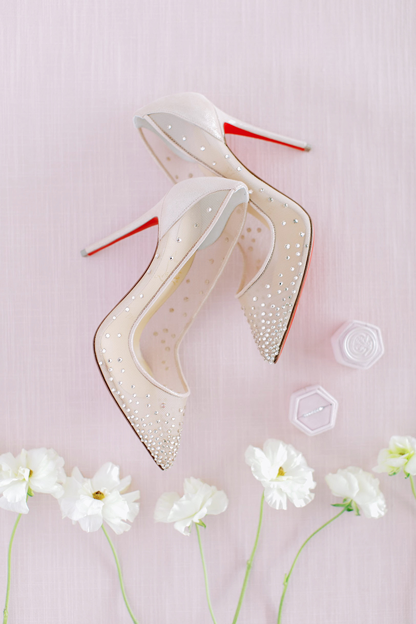 Flat Lay Photo of Nude Louboutin with gem accents surrounded by white flowers and a diamond wedding band