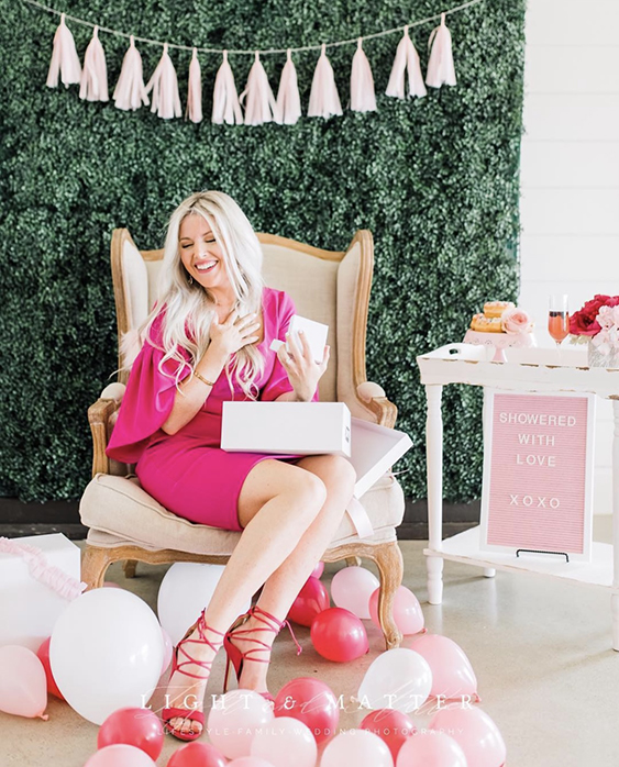 Blonde woman dressed in pink opening presents surrounded by balloons and treats, sitting in front of a boxwood wall.