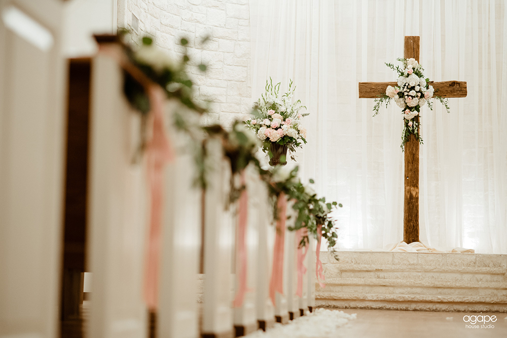 Barnwood Cross with floral centered on it in front of white drapery. Pews decorated with greenery, florals and pink ribbon.